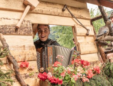 Eine lächelnde Person spielt ein Akkordeon in einer rustikalen Holzhütte, die mit roten Blumen und Grünzeug geschmückt ist.