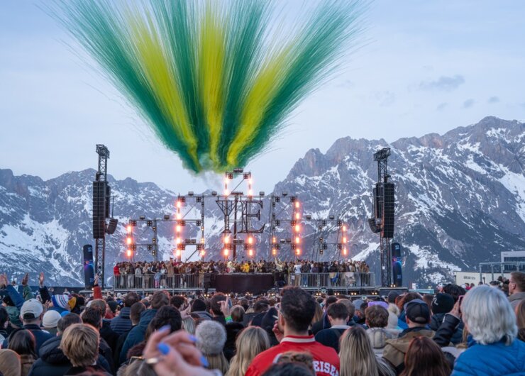 Eine Menschenmenge verfolgt ein Konzert auf einer Freilichtbühne mit farbigem Rauch und Bergen im Hintergrund.