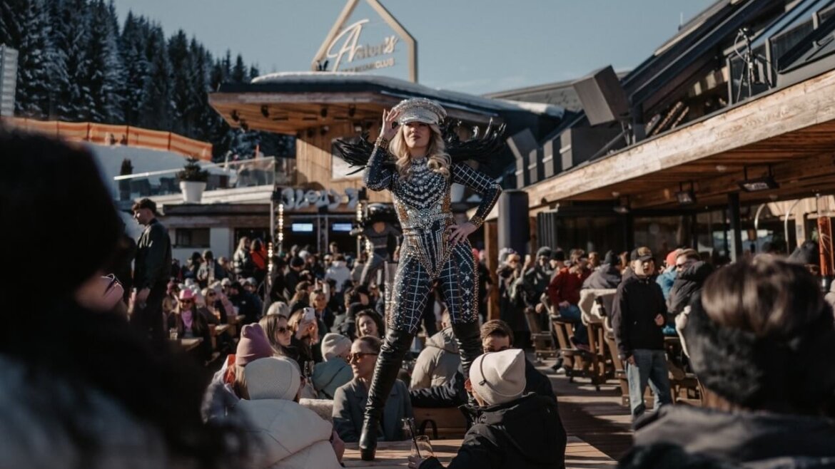 Ein Darsteller in einem silbernen Kostüm posiert auf einem Tisch in einem überfüllten Skigebiet mit verschneiten Bergen im Hintergrund. (vergrößerte Ansicht)