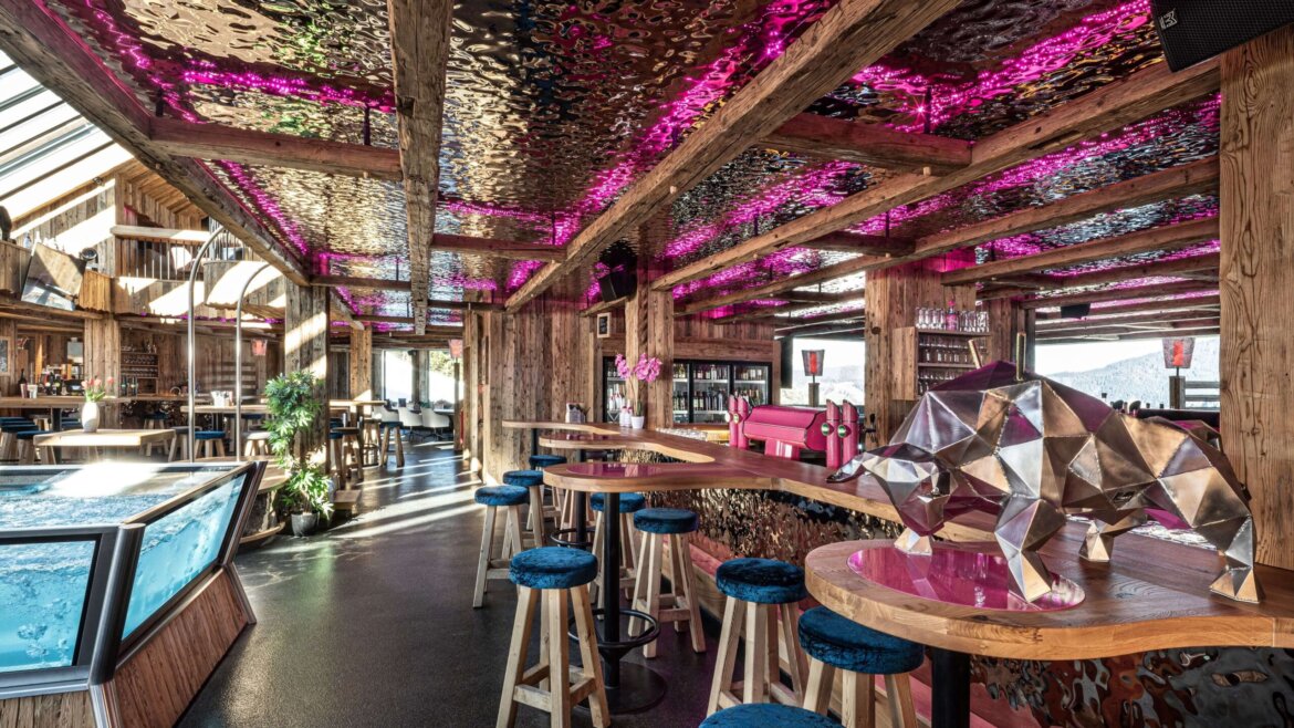 Eine rustikale Bar mit Holzbalken, rosa LED-Leuchten, blauen Hockern und einer geometrischen Bärenskulptur auf einem Tisch. (vergrößerte Ansicht)