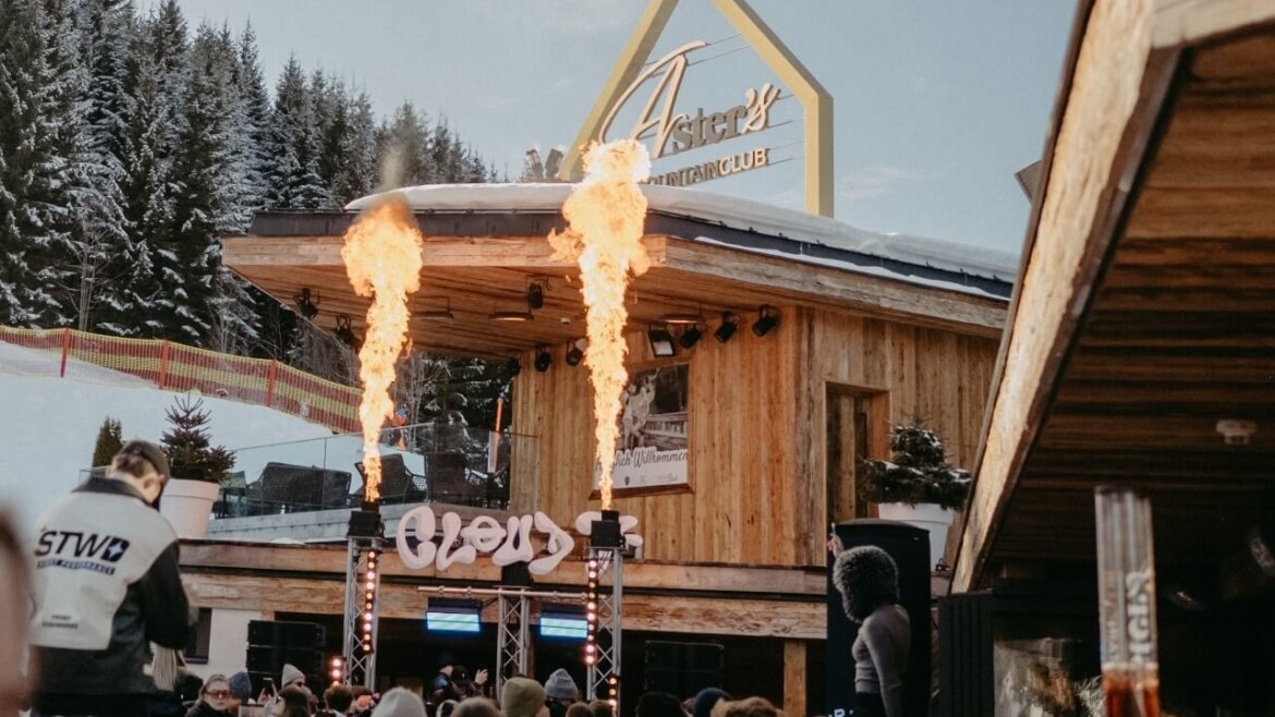 Eine Menschenmenge versammelt sich vor einer hölzernen Bergbar mit hochschießenden Feuerdüsen, umgeben von Schnee und Bäumen. (vergrößerte Ansicht)
