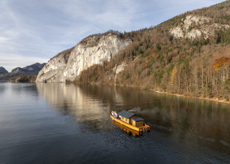 Ein Holzboot schwimmt auf einem ruhigen See in der Nähe von bewaldeten Hügeln und einer großen weißen Felsklippe.