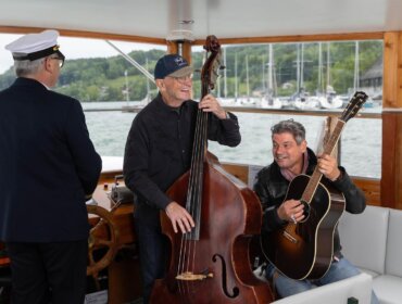 Schiffsfahrten_mit_Live-Musik_an_den_Trumer_Seen ©Seeham Zwei Männer spielen Gitarre und Kontrabass auf einem Boot, während ein Kapitän am Steuer steht. Segelboote im Hintergrund.