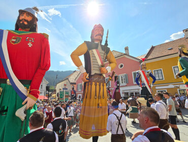 Menschen ziehen mit riesigen traditionellen Puppen in bunten Kostümen unter einem sonnigen Himmel auf einem europäischen Stadtplatz umher.