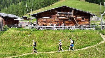 Drei Wanderer gehen auf einem grasbewachsenen Weg vor einer hölzernen Berghütte, vor der Menschen sitzen.