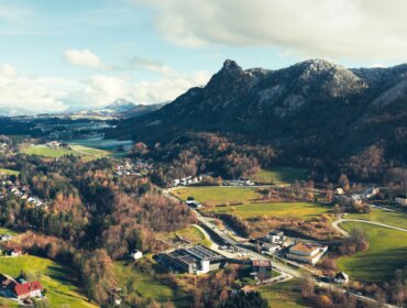 Luftaufnahme eines Dorfes mit verstreuten Häusern, grünen Feldern und bewaldeten Bergen unter einem teilweise bewölkten Himmel.