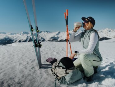 Mindful Escapes im SalzburgerLand: Sandra Lahnsteiner-Wagner Ein Skifahrer kniet im Schnee und trinkt aus einer Flasche, während im Hintergrund Berge und Skiausrüstung zu sehen sind.