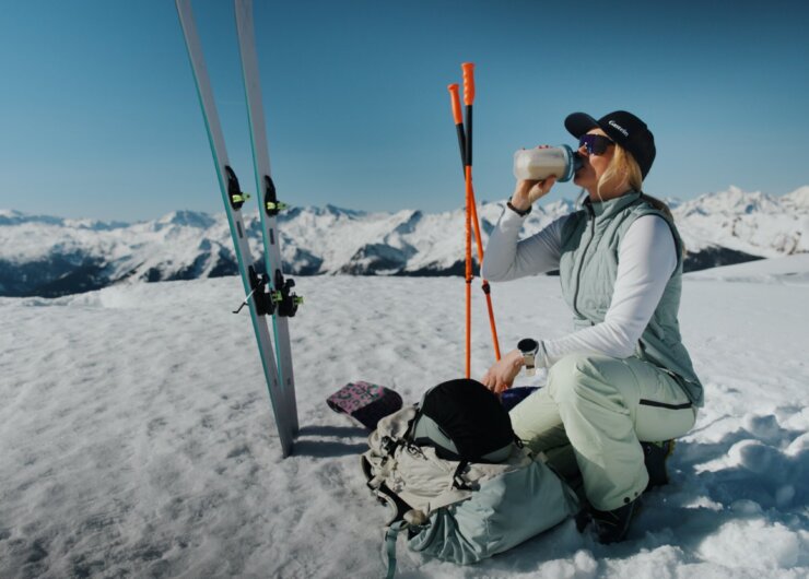 Ein Skifahrer kniet im Schnee und trinkt aus einer Flasche, während im Hintergrund Berge und Skiausrüstung zu sehen sind.