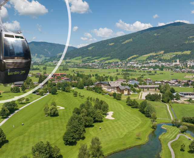 An einem sonnigen Tag gleitet eine Seilbahn über einen grünen Golfplatz mit Bergen und einem Dorf im Hintergrund.
