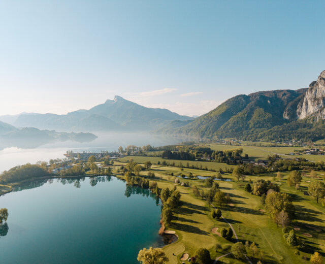 Luftaufnahme eines ruhigen Sees, umgeben von grünen Feldern, Bäumen und entfernten Bergen unter einem klaren Himmel.