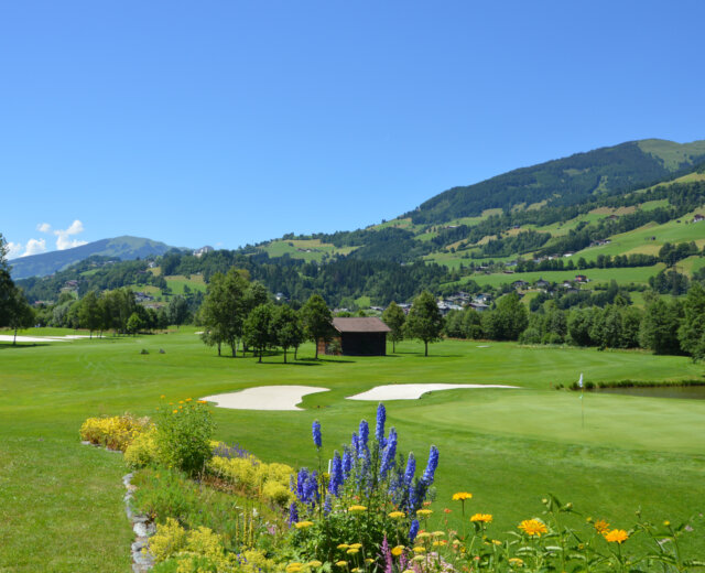 Ein Golfplatz mit Sandbunkern, grünen Hügeln, Blumen und Bergen unter einem strahlend blauen Himmel.