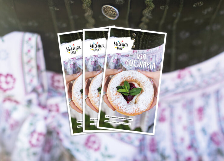 Drei Broschüren mit einem Gebäck auf dem Umschlag, die vor eine Person in traditioneller Kleidung gehalten werden.