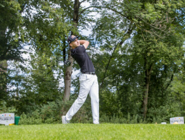 Ein Golfer im Schwung auf einem grünen Platz, umgeben von Bäumen und blauem Himmel.