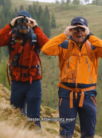 Zwei Personen in Outdoor-Kleidung beobachten mit einem Fernglas die Landschaft auf dem Salzburger Almenweg. Text: Der beste Arbeitsplatz aller Zeiten!.