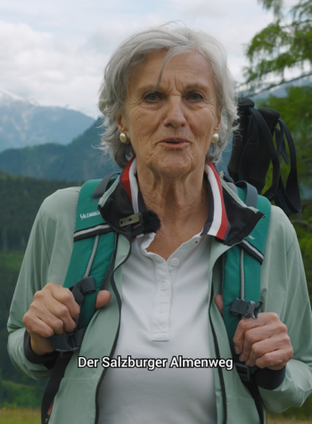 Eva Mayr, die Impulsgeberin des Salzburger Almenweges und einem Rucksack steht im Freien, hinter ihr erheben sich die Berge auf dem Salzburger Almenweg.