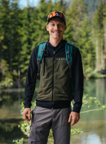 Jan Hörl steht lächelnd in Outdoor-Kleidung und mit Rucksack am Almsee in Filzmoos entlang des Salzburger Almenweges
