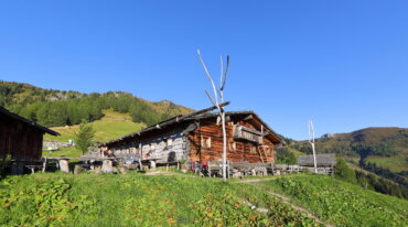 Eine rustikale Holzhütte steht auf einem grasbewachsenen Hügel unter einem strahlend blauen Himmel, umgeben von Bergen und Bäumen.