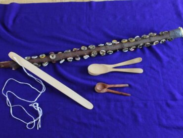 Vier hölzerne Musikinstrumente, darunter ein Stock mit Muscheln, auf einem violetten Stoffhintergrund.