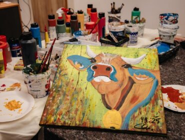 Eine bunte Kuh malt auf einem Tisch, umgeben von Farben, Pinseln und Kunstzubehör.