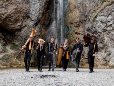 ©Autländisch_Fmily Sechs Musiker mit verschiedenen Instrumenten stehen vor einem felsigen Wasserfall und lächeln in die Kamera.