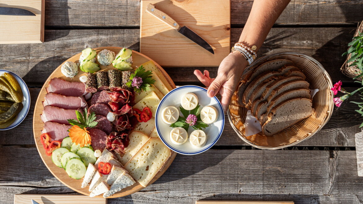 Eine Hand hält einen Teller mit Butterscheiben zwischen einem Brotkorb und einem Jausenbrett – ideal für den Almsommer im Grossarltal. (vergrößerte Ansicht)