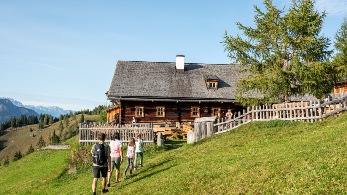 Vier Menschen wandern auf einem grasbewachsenen Hügel auf ein Holzhaus im Nationalpark Hohe Tauern zu, hinter dem sich die Berge erheben.