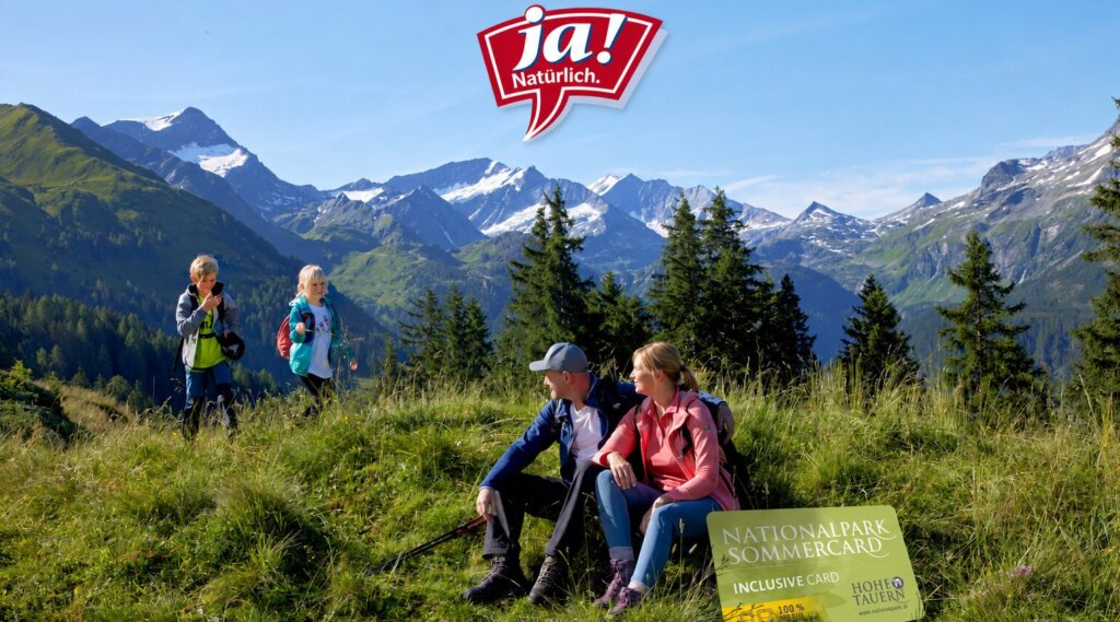 Eine Familie wandert in den grünen Bergen mit schneebedeckten Gipfeln und sitzt in der Nähe eines Schildes für den Nationalpark Sommercard.
