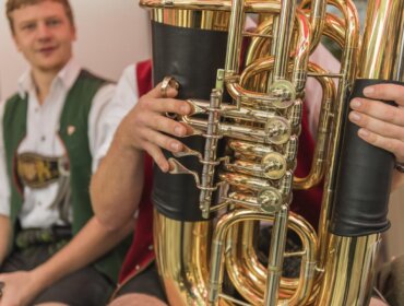 Mann in traditioneller Kleidung spielt ein glänzendes Blechblasinstrument, ein anderer Mann im Hintergrund, leicht unscharf.