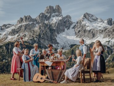 Eine Gruppe in traditioneller Tracht spielt im Freien vor schneebedeckten Bergen und einem Holztisch auf verschiedenen Instrumenten.
