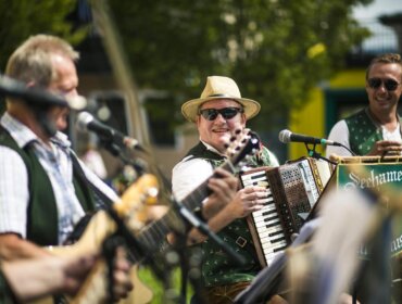 @Seeham Vier Männer in grünen Westen spielen Instrumente und singen im Freien, lächeln und genießen den sonnigen Tag.