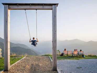 dorfgastein-Himmelsschaukel Ein Kind schaukelt auf der Himmelsschaukel am Fulseck in Dorfgastein, während vier Personen auf Bänken sitzen und den Blick auf die Berge bei Sonnenuntergang genießen.