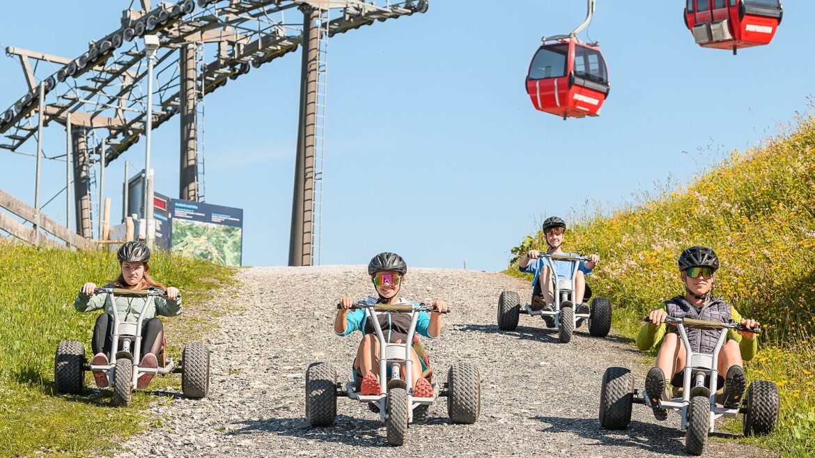 © Dorfgasteiner Bergbahnen AG, Mountaincart An einem sonnigen Tag fahren vier Kinder mit Mountaincarts auf einem Schotterweg vor den Dorfgasteiner Bergbahnen bergab. (vergrößerte Ansicht)