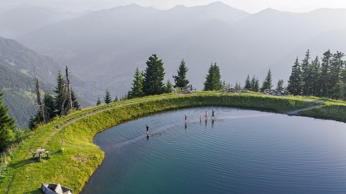 © Dorfgasteiner Bergbahnen AG, Spiegelsee Menschen stehen im Spiegelsee in Dorfgastein, umgeben von Bäumen, im Hintergrund sind ferne Berge zu sehen. (vergrößerte Ansicht)