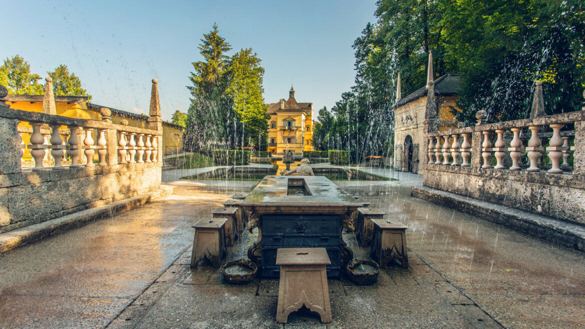 (c) SalzburgerLand Tourismus, Eva trifft., Wasserspiele Salzburg Ein Innenhof mit einem Steinbrunnen, Wasserstrahlen, üppigem Grün und der gelben Villa „Salzschleife“ im Hintergrund. (vergrößerte Ansicht)