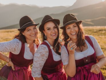 Hollerstauden Titelfoto Drei Frauen in einheitlichen roten Kleidern und Hüten lächeln im Freien, hinter ihnen sind die österreichischen Berge und der Sonnenschein zu sehen.