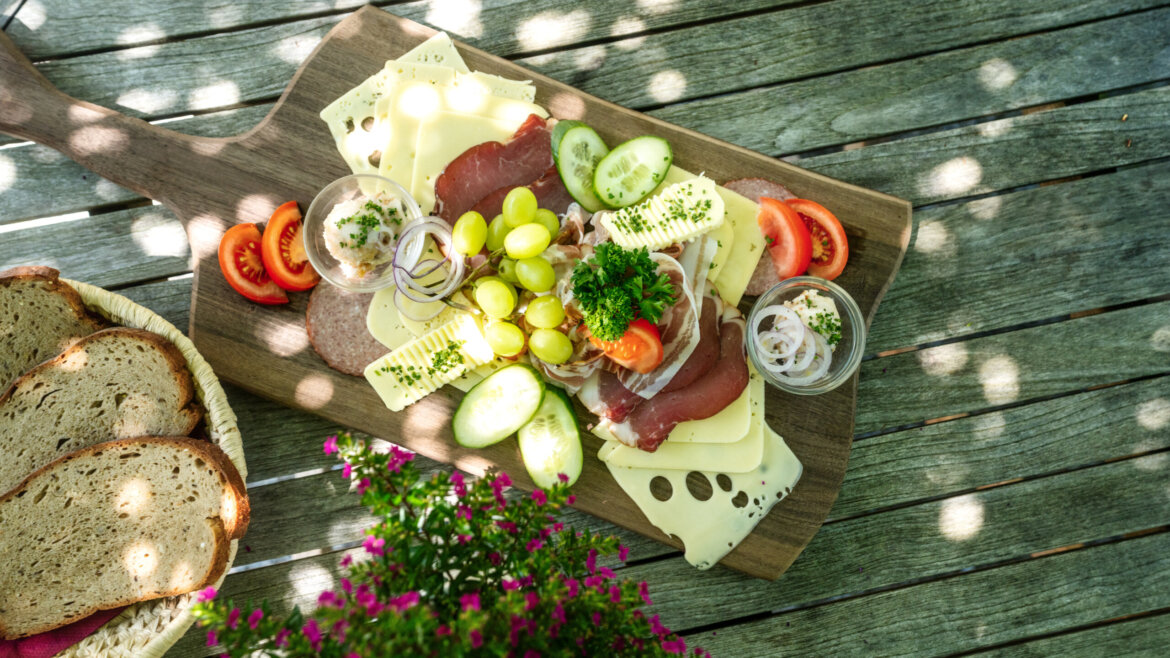 Talwirt_Thema Genuss (1) © Daniel Kogler – Ferienregion Nationalpark Hohe Tauern Eine Charcuterie-Platte mit Käse, Fleisch, Weintrauben, Gemüse und Brot auf einem Holztisch im fleckigen Sonnenlicht. (vergrößerte Ansicht)