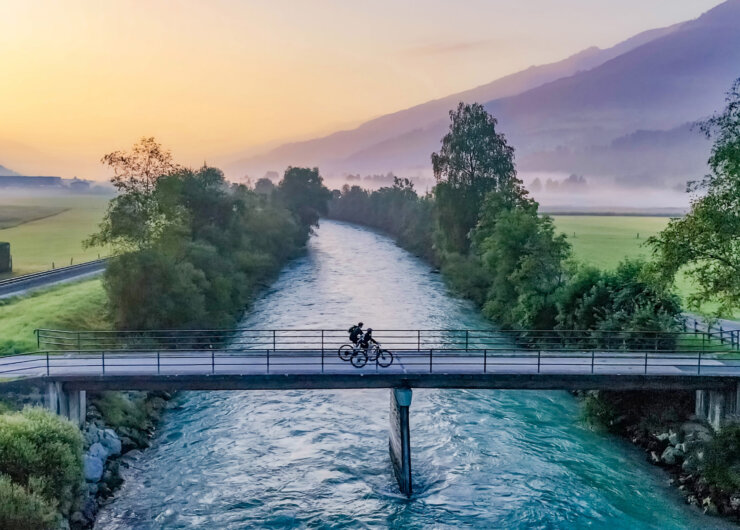Tauernradweg_Salzachbrücke (c) Ferienregion Nationalpark Hohe Tauern – Agentur Lichtfarben Zwei Radfahrer überqueren bei Sonnenaufgang eine schmale Brücke über einen Fluss, im Hintergrund sind nebelverhangene Berge zu sehen.