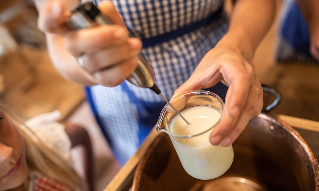 © SalzburgerLand Tourismus, Michael Groessinger, Bio-Kosmetik Eine Person, die eine blau karierte Schürze trägt, verrührt mit einem Handmilchaufschäumer Milch in einem Glasbecher. (vergrößerte Ansicht)