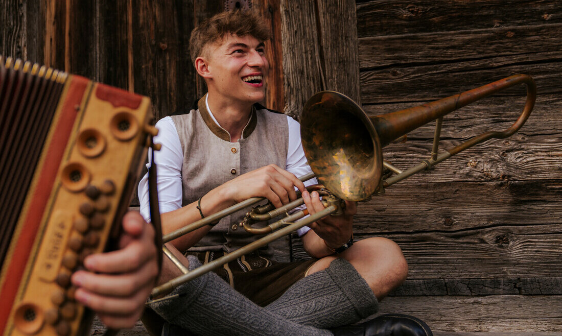© SalzburgerLand Tourismus, Lisa Eiersebner, Musik im Bauernherbst Ein lächelnder junger Mann in traditioneller Kleidung sitzt an einer Holzwand und hält ein Blechblasinstrument in der Hand. Ein Akkordeon ist zu sehen. (vergrößerte Ansicht)