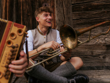 Musik im Bauernherbst Ein lächelnder junger Mann in traditioneller Kleidung sitzt an einer Holzwand und hält eine Trompete in der Hand. Eine Zieharmonika ist zu sehen.