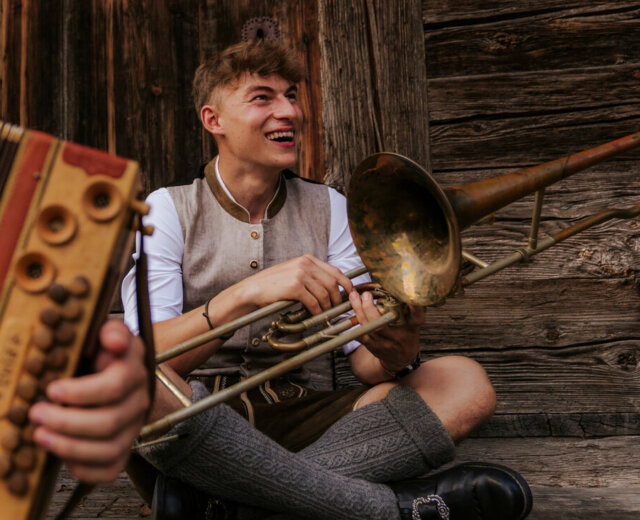 Musik im Bauernherbst Ein lächelnder junger Mann in traditioneller Kleidung sitzt an einer Holzwand und hält eine Trompete in der Hand. Eine Zieharmonika ist zu sehen.