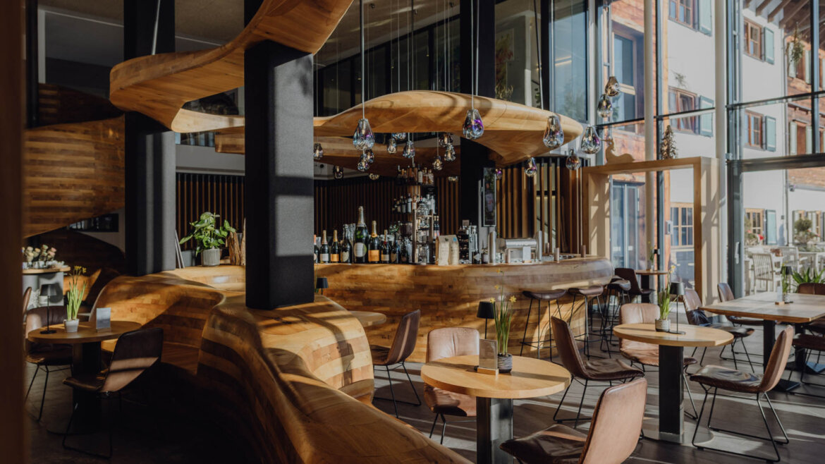 Bar Freiraum © Puradies by Lukas Reschreiter Ein modernes Café mit Holzmöbeln, einer geschwungenen Theke, Hängelampen und großen Fenstern, durch die Sonnenlicht hereinströmt. (vergrößerte Ansicht)