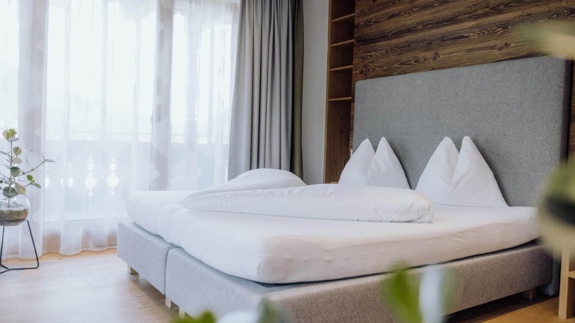 Zimmer © Puradies by creating click Modernes Schlafzimmer mit einem Doppelbett, weißer Bettwäsche, Holzakzenten, einem großen Fenster und Zimmerpflanzen. (vergrößerte Ansicht)