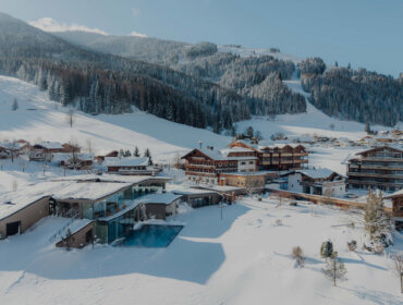Gesamtanlage Hotel Außen Winter 2026 © Puradies by Lukas Reschreiter Ein verschneites Dorf in einem Bergtal mit Chalets, immergrünen Bäumen und einem Pool unter strahlend blauem Himmel.