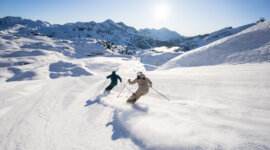 Zwei Skifahrer gleiten auf einem präparierten Hang durch den Neuschnee, während über ihnen ein strahlend blauer Himmel und in der Ferne Berggipfel zu sehen sind.