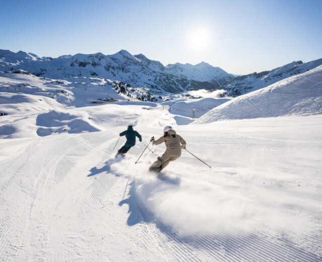 Zwei Skifahrer gleiten auf einem präparierten Hang durch den Neuschnee, während über ihnen ein strahlend blauer Himmel und in der Ferne Berggipfel zu sehen sind.