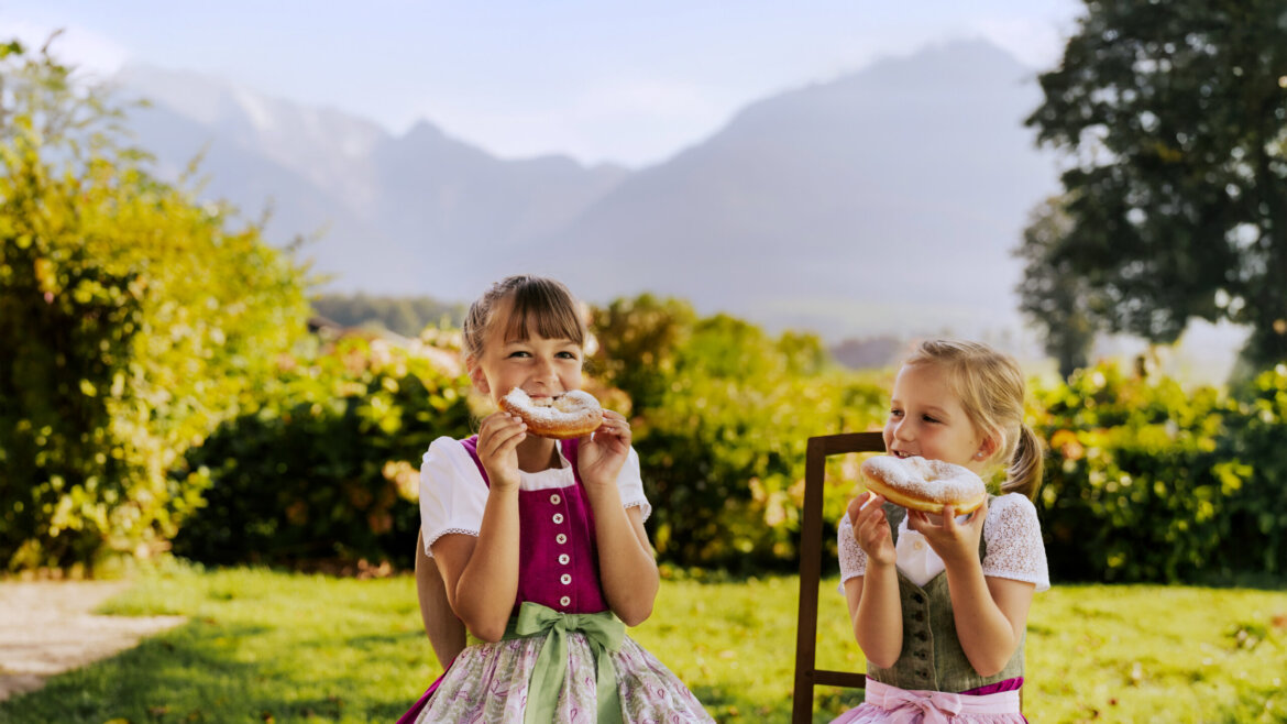 © SalzburgerLand Tourismus, Lisa Eiersebner, Bauernkrapfen Zwei junge Mädchen in traditionellen österreichischen Trachten essen draußen inmitten von Bergen und grüner Natur Gebäck. (vergrößerte Ansicht)
