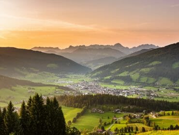 A scenic valley at sunset with Altenmarkt-Zauchensee, green fields, forests, and distant mountain ranges.