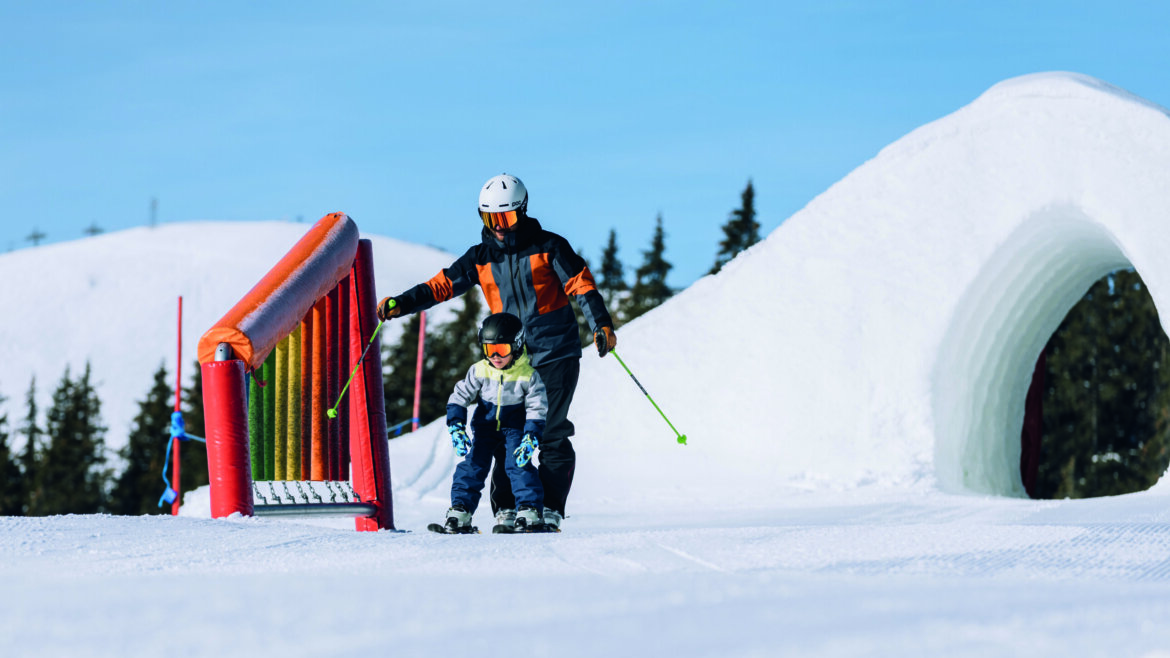 Family in Skicircus Saalbach-Hinterglemm-c-Mia-Knoll An adult teaches a child to ski on a snowy Saalbach Hinterglemm slope with obstacles and a tunnel behind. (Enlarged view)