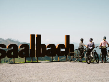 Three cyclists with bikes stand by a large Saalbach Hinterglemm sign, overlooking mountains on a clear day.